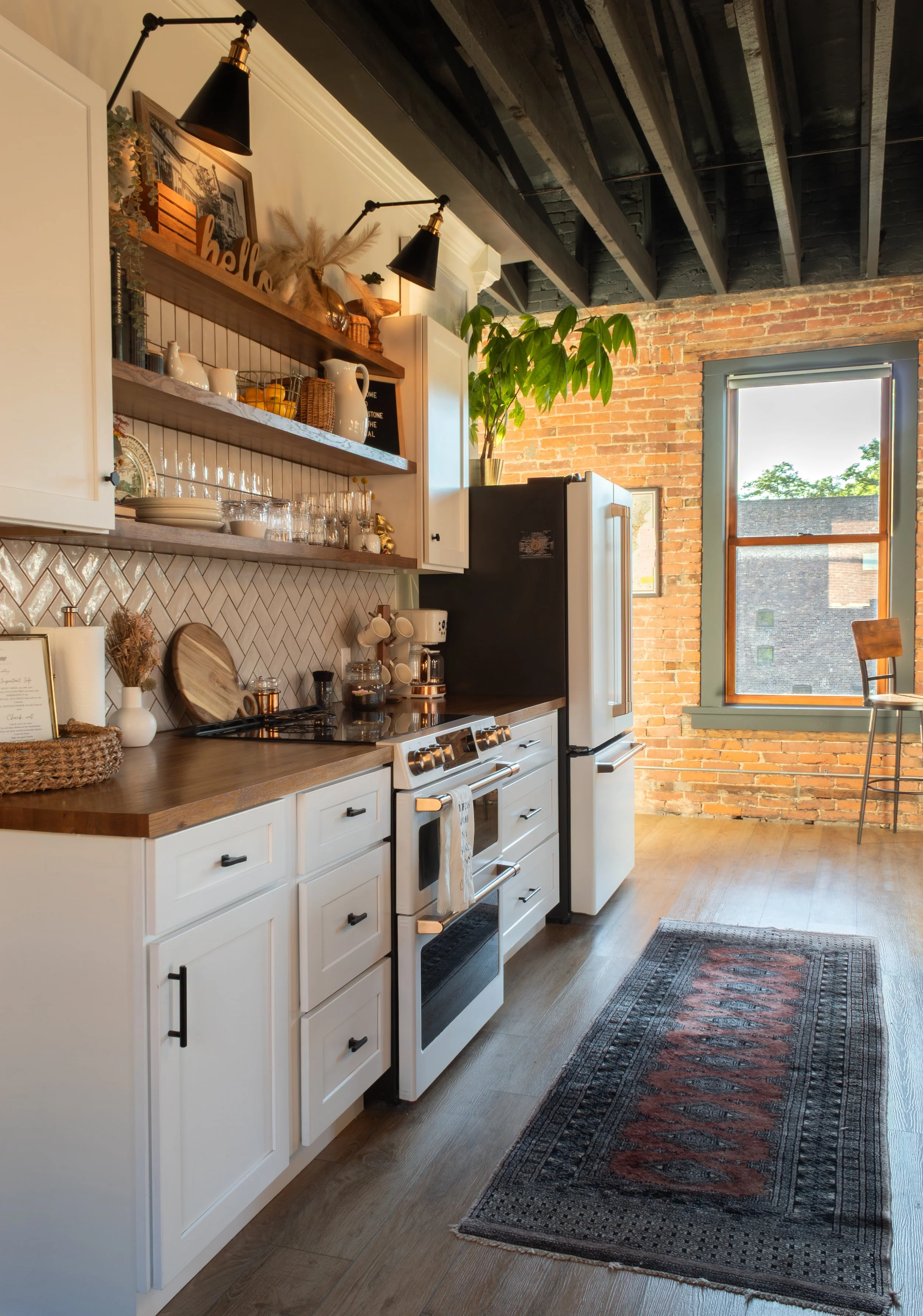 A modern kitchen with white cabinetry, a wooden countertop, open shelves with dishes and decorative items, a black refrigerator, a window with a view of a building and trees, brick wall, and a colorful area rug on wooden flooring.