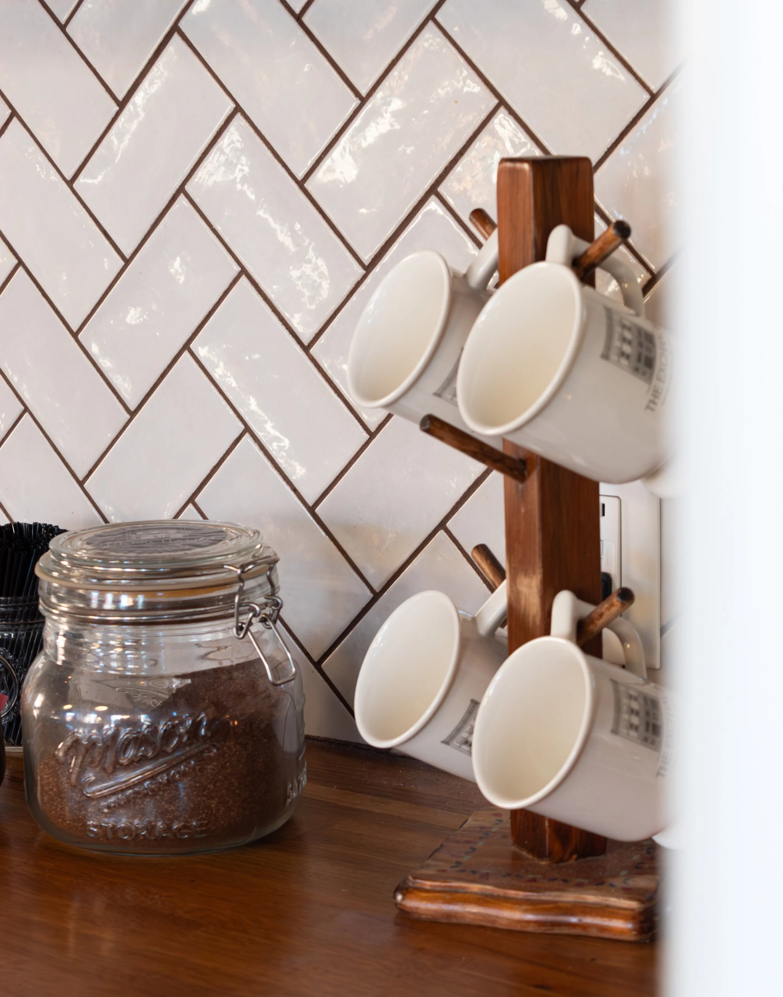 A kitchen counter with a glass jar of ground coffee, stacked glass jars, and a wooden mug rack with three white coffee mugs. The wall behind has white, rectangular tiles with dark grout.