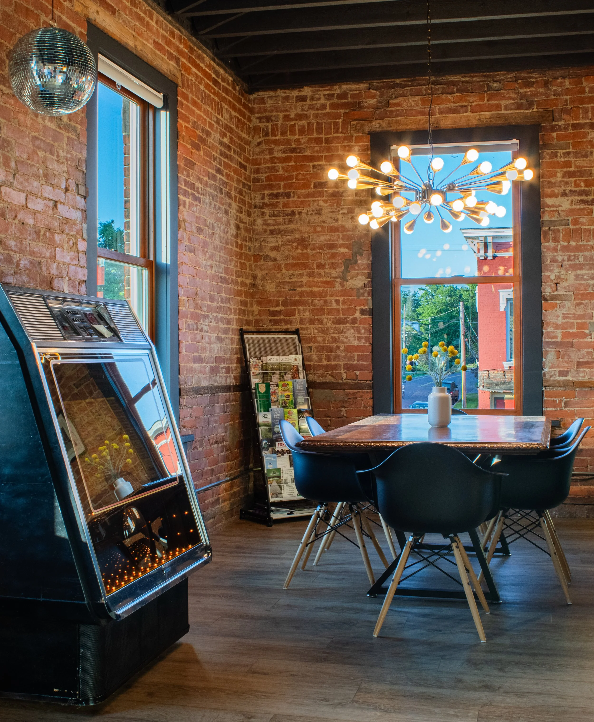 Interior of a restaurant or cafe with exposed brick walls, a window with black trim, a modern chandelier with multiple bulbs, a table with black chairs, and a small white vase with yellow flowers.