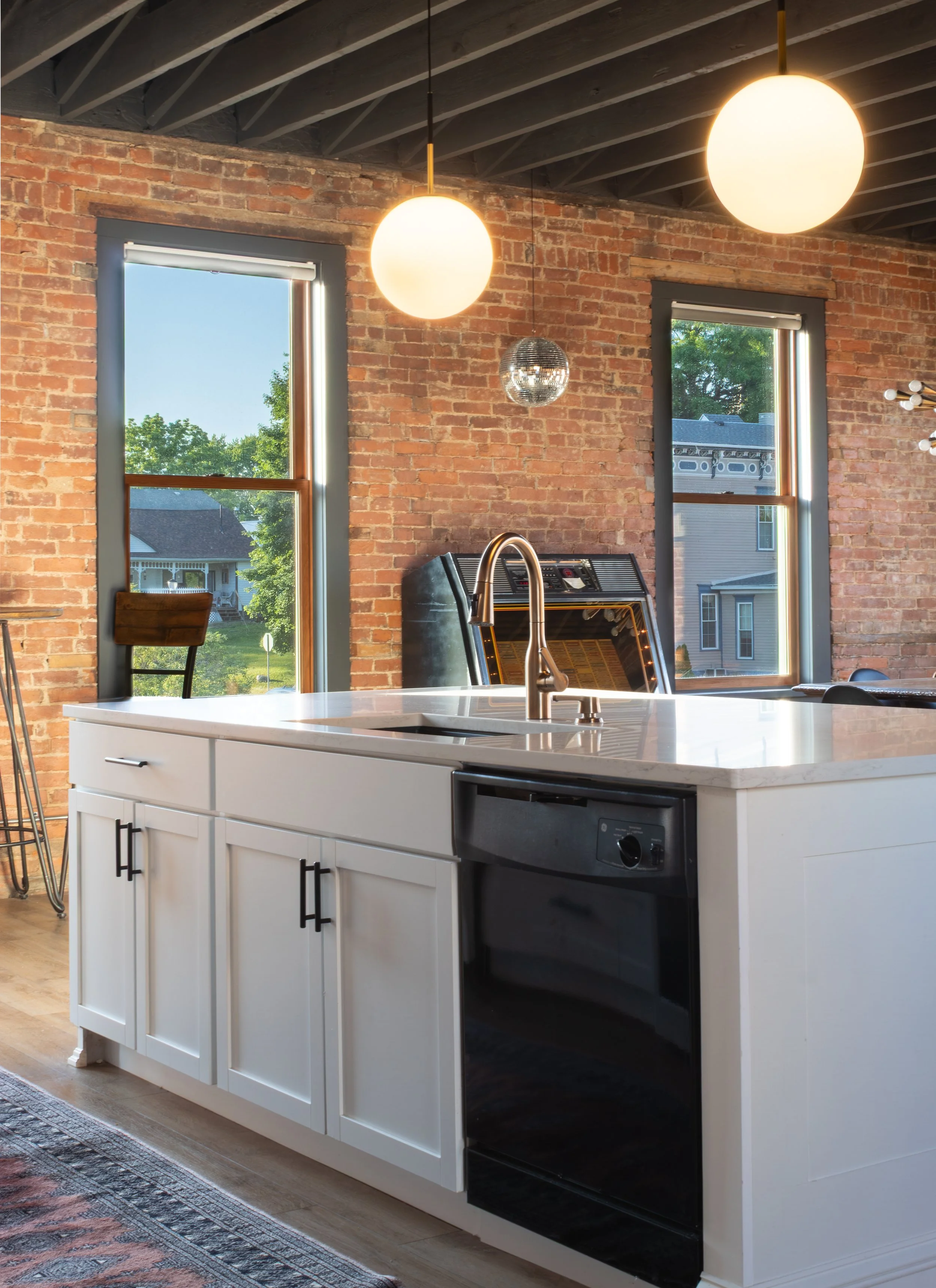 Modern kitchen with white island counter, black trash bin, exposed brick walls, large windows with views of trees and neighboring houses, and ceiling-mounted round light fixtures.