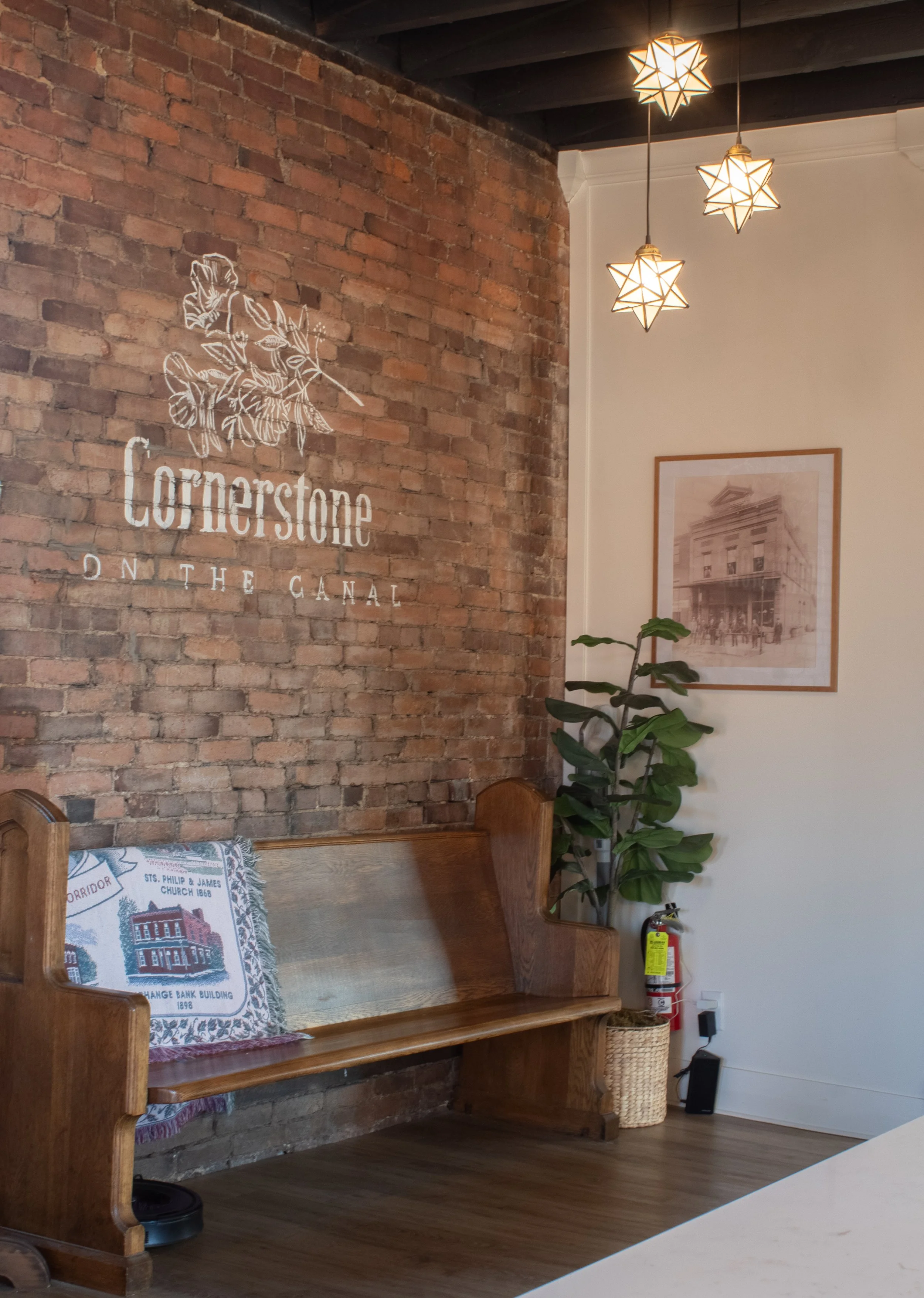 Interior of a cozy cafe or waiting area with a brick wall that features the logo 'Cornerstone on the Canal' with a floral illustration. There's a wooden bench with embroidered pillows referencing historical buildings and churches, a green plant, fram