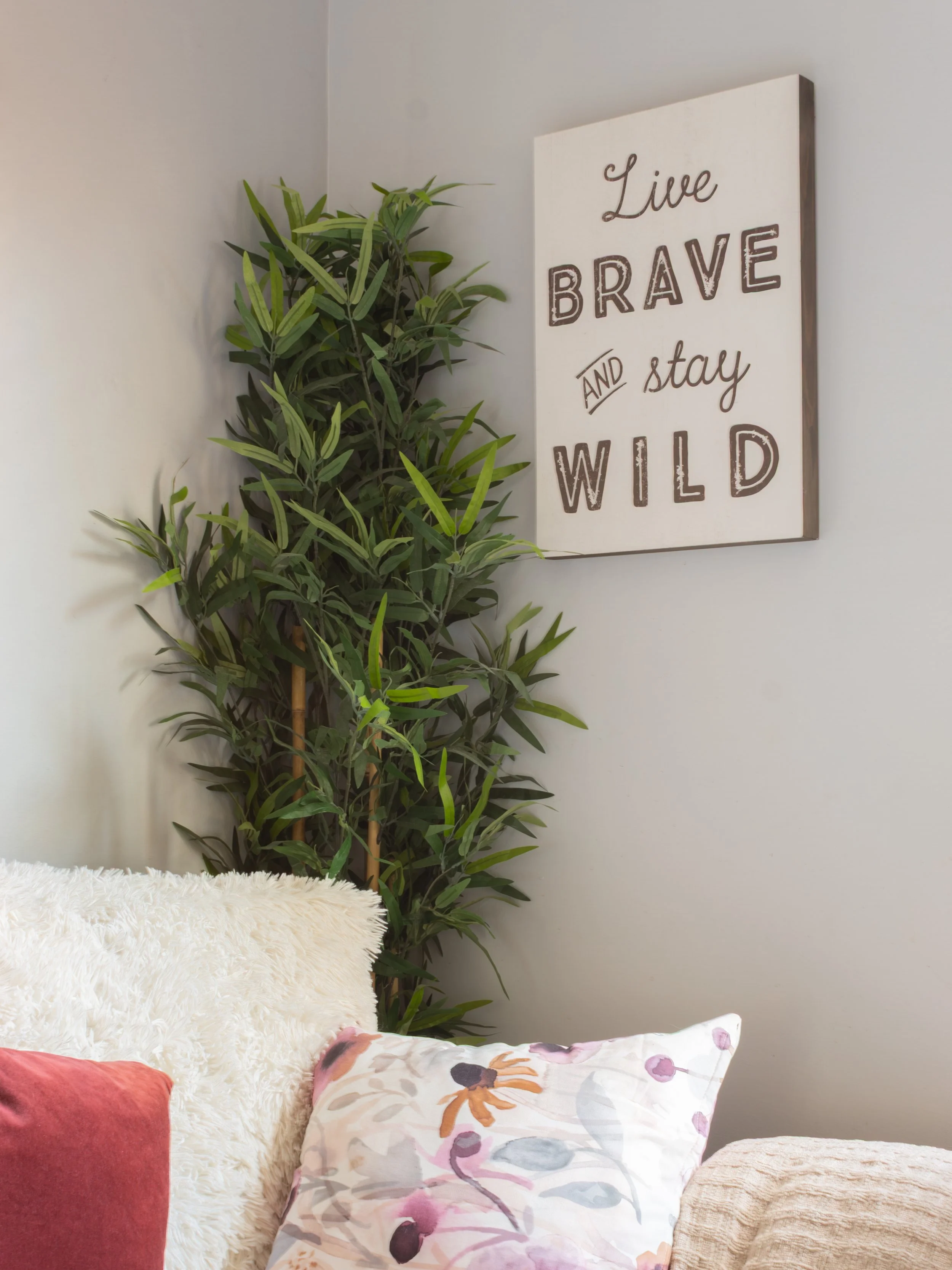 A cozy corner with decorative pillows, a fluffy white throw, a large leafy green plant, and a wall sign that reads 'Live brave and stay wild' in cursive and bold lettering.