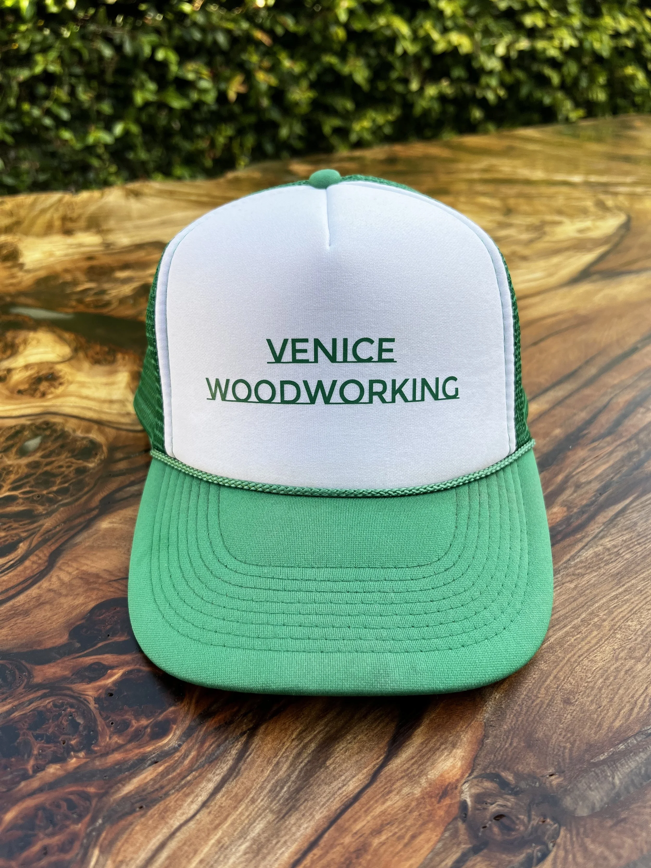 A baseball cap with a green brim and white front panel. The front panel has the text "VENICE WOODWORKING" printed on it. The cap is placed on a wooden surface outdoors with a green hedge in the background.
