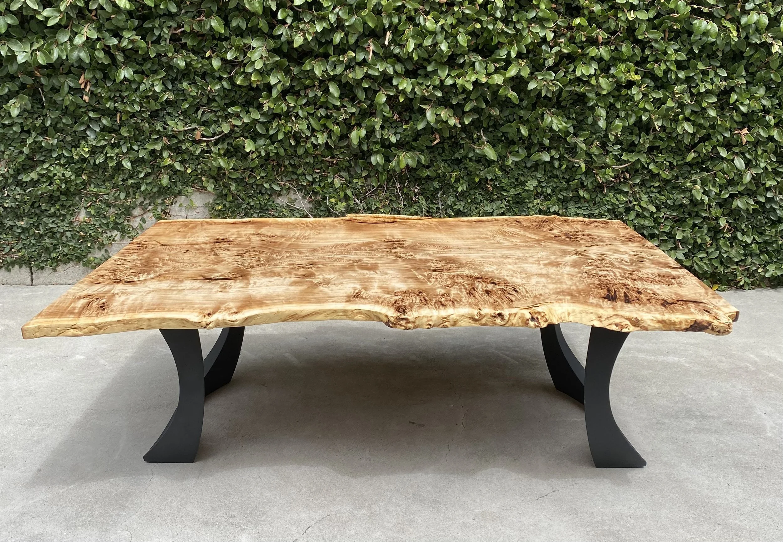 A wooden table with a live edge design, supported by black metal legs, placed outdoors in front of a green hedge.