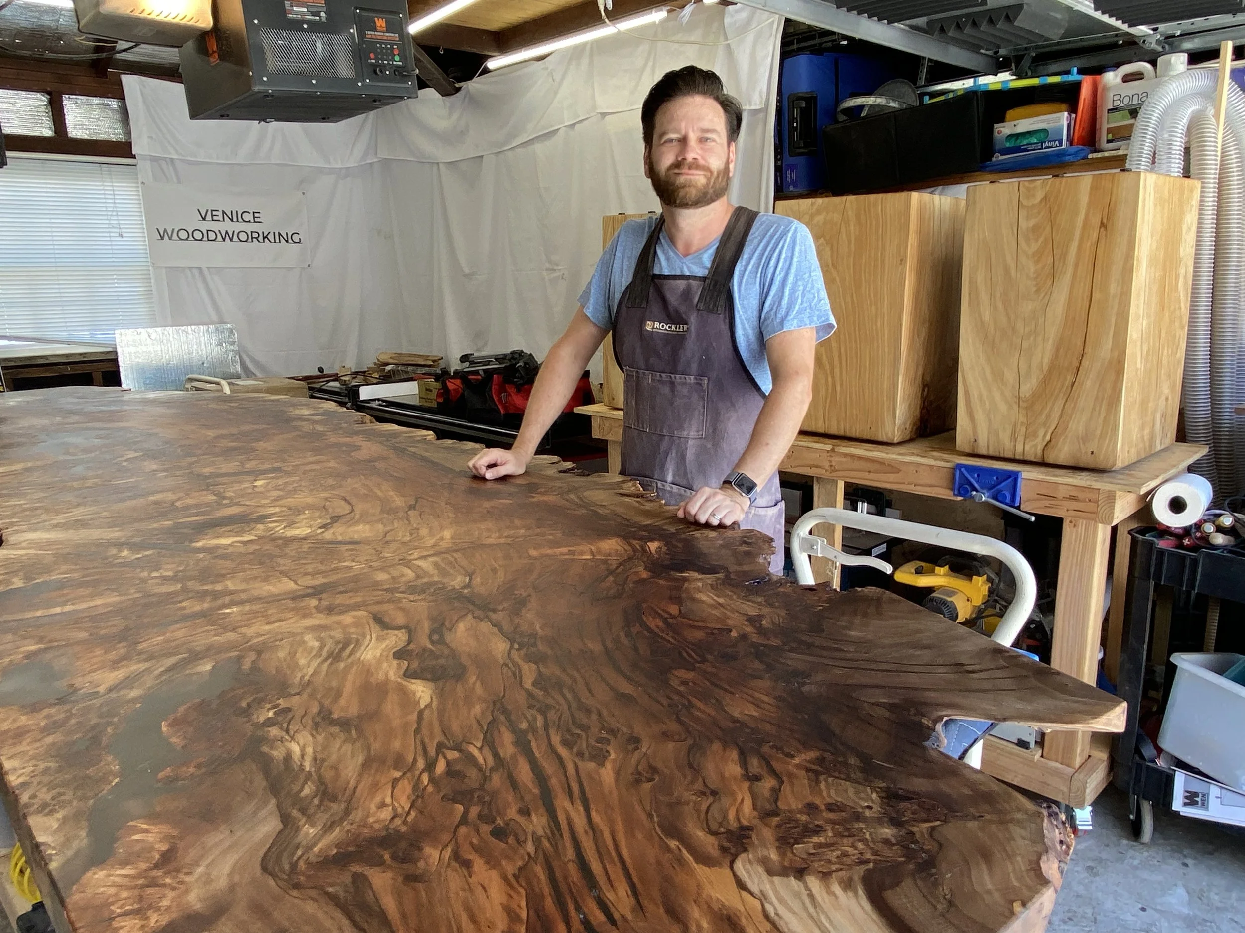 A man in a workshop stands behind a large, polished wooden table with intricate grain patterns.  The workshop has shelves filled with various tools and supplies, and a sign on the wall reads "Venice Woodworking." The space is well-lit with natural and artificial light.