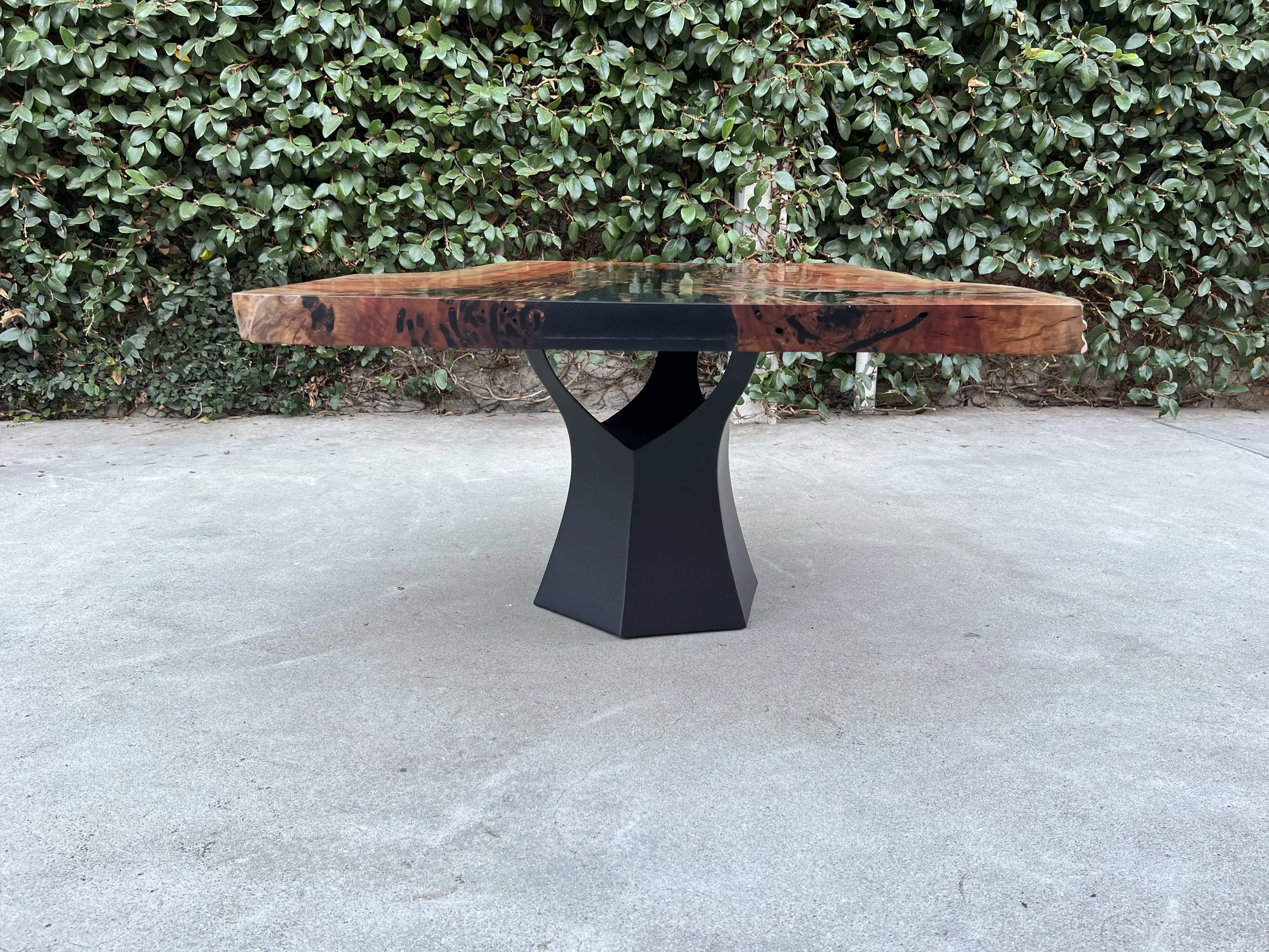 A round wooden table with a dark metal base placed outdoors on a concrete surface, with a green leafy hedge in the background.