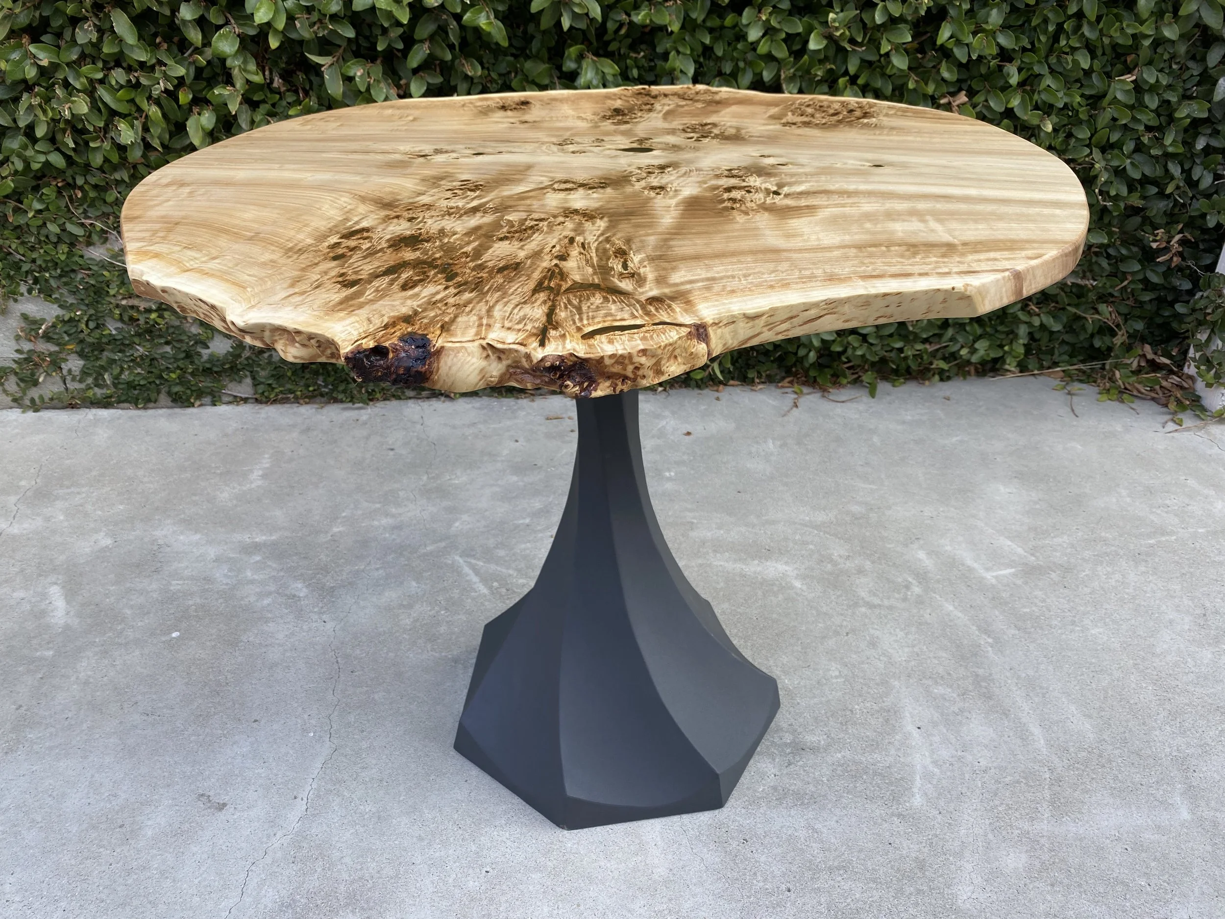 A wooden table with a natural, live edge finish, placed outside in front of green shrubbery and a concrete surface.