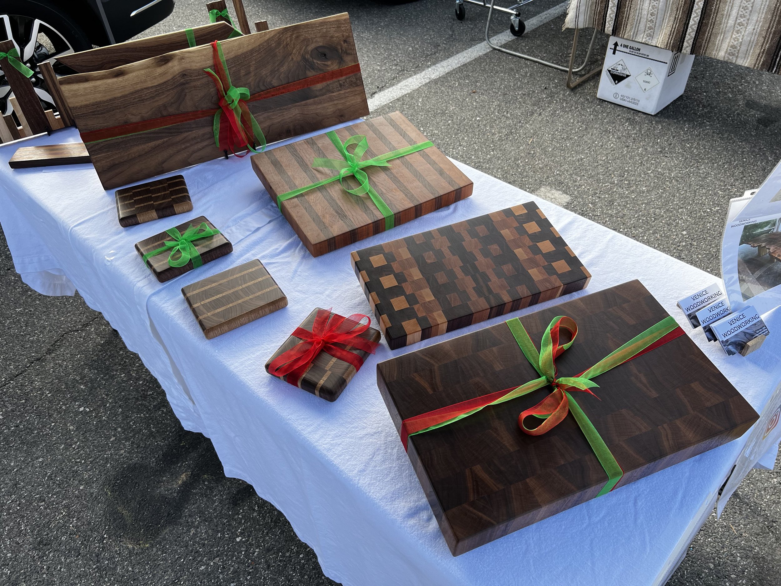 Various wooden cutting boards and serving platters with different wood grain patterns, decorated with colorful ribbons, are displayed on a table covered with a white tablecloth at an outdoor market.