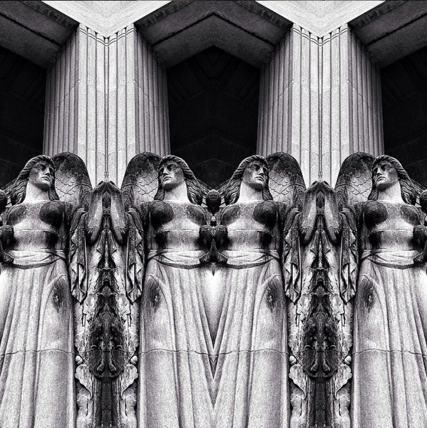 Black and white photo of a symmetrical architectural detail featuring three large stone angel sculptures with wings, positioned behind tall fluted columns.