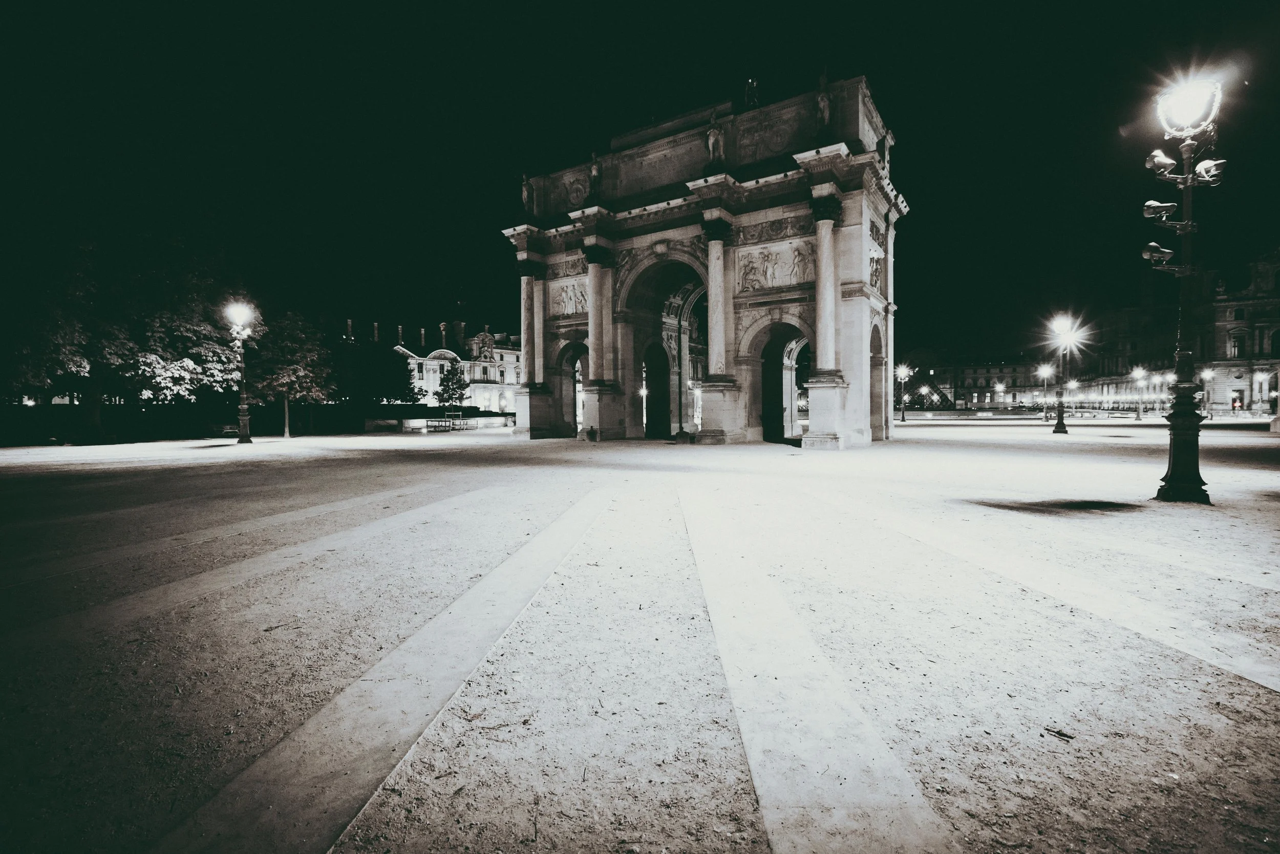 Nighttime view of a historic archway monument illuminated by streetlights, with a clear, empty street in front and city buildings in the background.