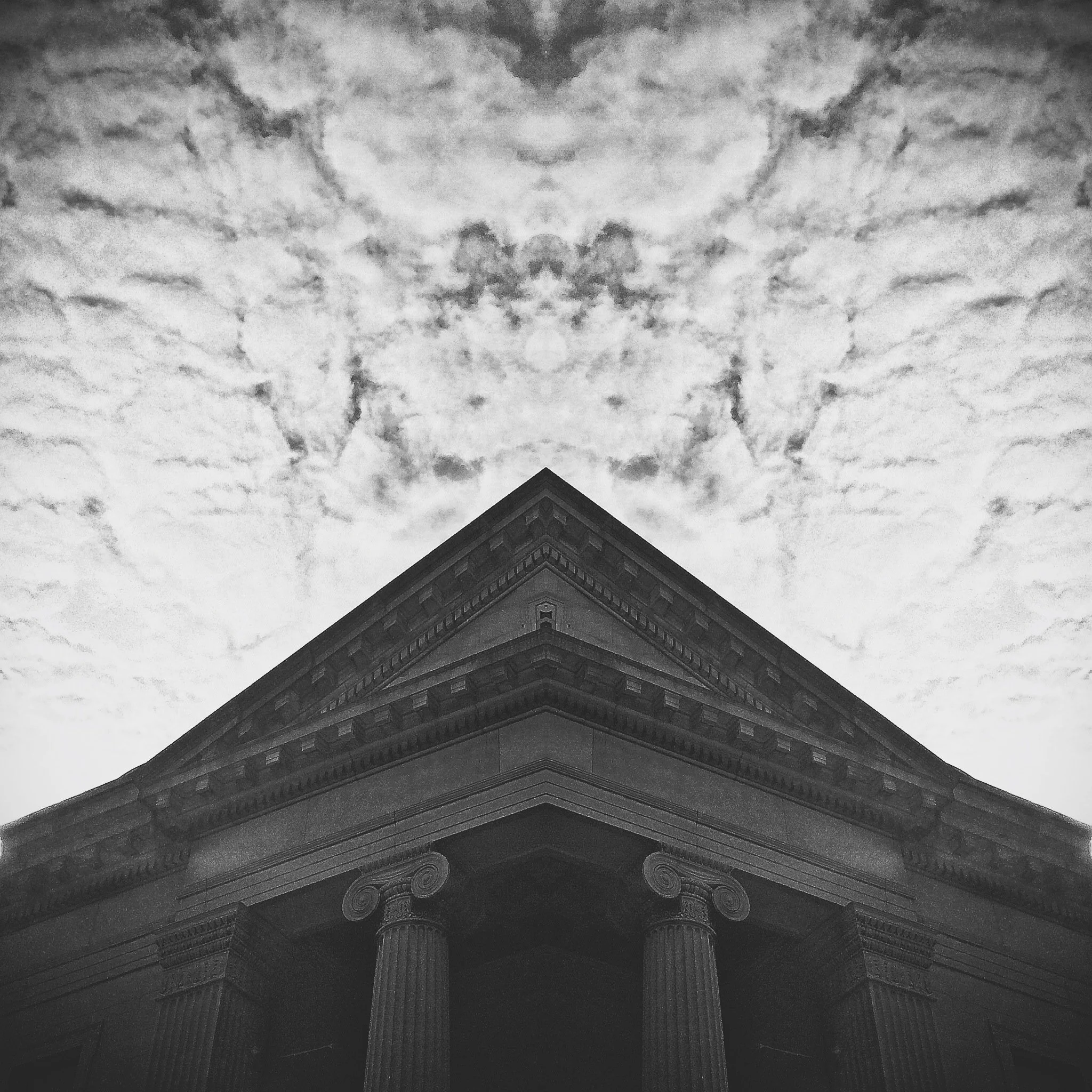 Black and white photo of an ornate building with detailed architectural elements and a dark roof, viewed from below against a cloudy sky.