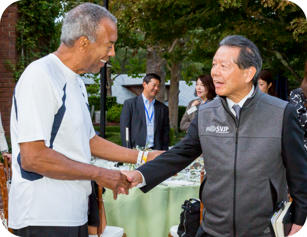 Two men shaking hands at an outdoor event, with several people standing in the background near a table set for a meal under trees.