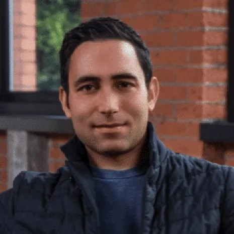 A man with short dark hair and light skin sitting outdoors on a porch with brick walls, wearing a dark vest and a blue shirt, smiling