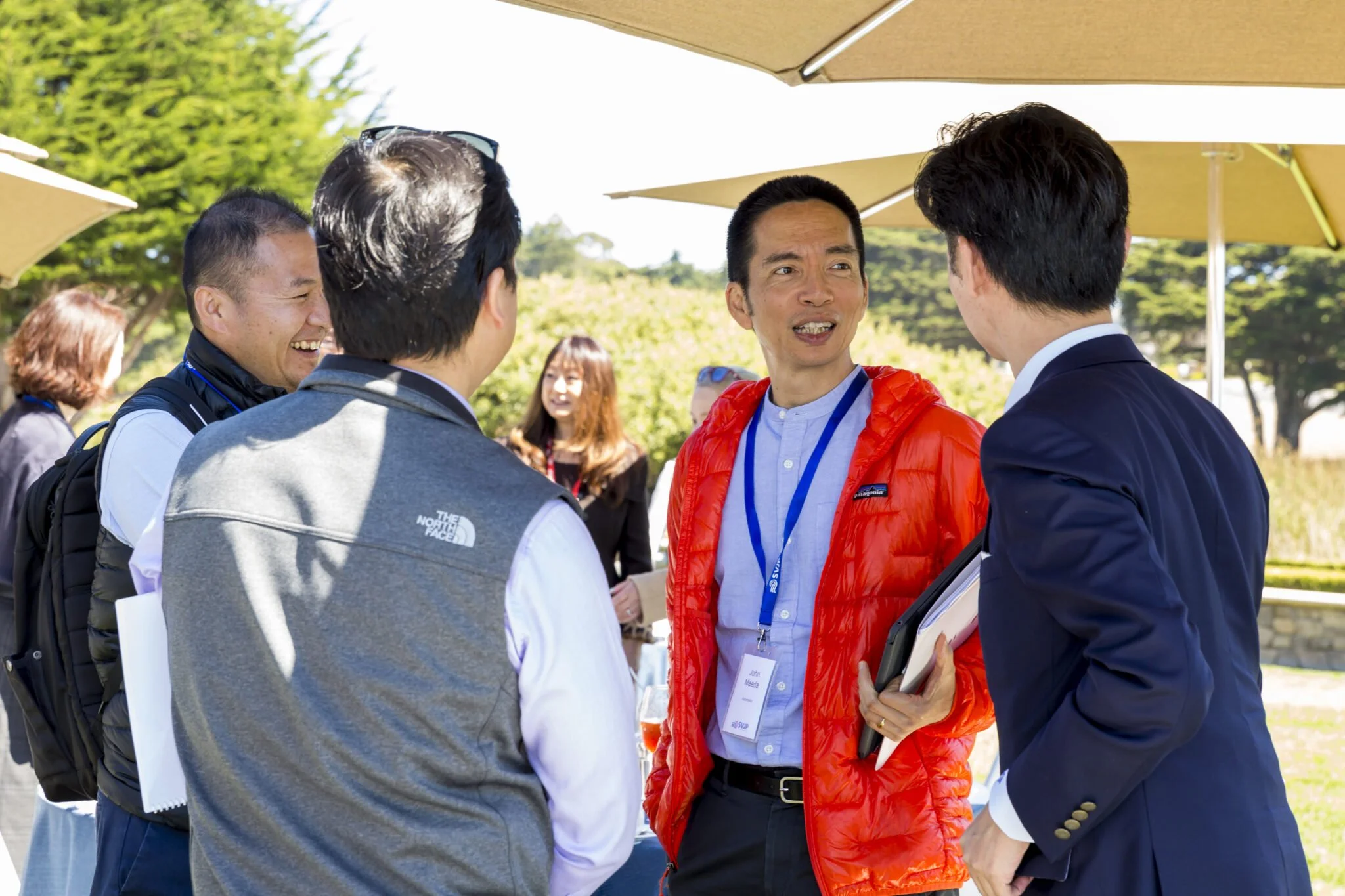 People engaged in conversation outdoors under umbrella tents, with trees and clear sky in background.