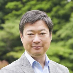 Man with short dark hair wearing a light gray blazer over a white shirt outdoors with green foliage in the background.