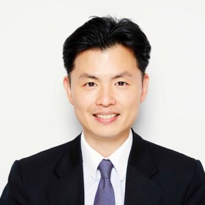 Professional headshot of a man in a black suit, white shirt, and purple tie, smiling against a plain white background.