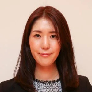 Portrait of a woman with shoulder-length brown hair wearing a black blazer and patterned blouse, smiling against a light background.
