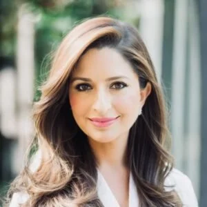 A woman with long, wavy brown hair, smiling, outdoors with green foliage in the background.