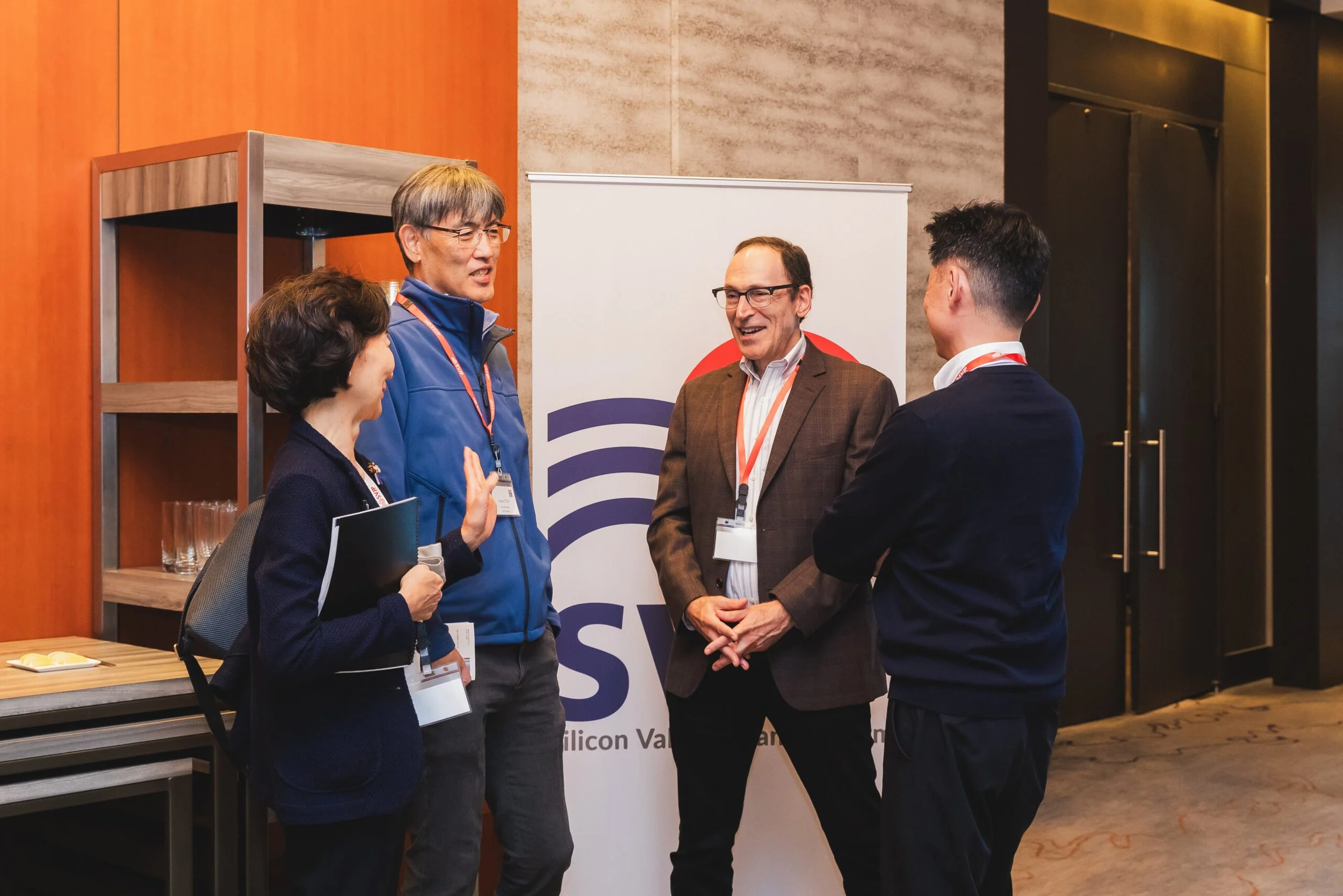 Four people engaged in conversation at a professional event, standing near a banner that reads 'Silicon Valley.' They are wearing conference badges and business attire, smiling and gesturing as they speak.