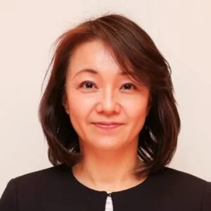 A woman with shoulder-length brown hair wearing a black top against a plain light-colored background.