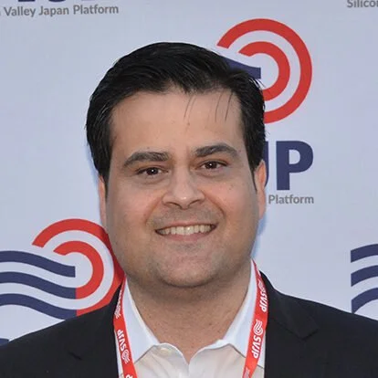 A man with dark hair in a suit smiling at the camera, standing in front of a backdrop with red and blue logo designs.