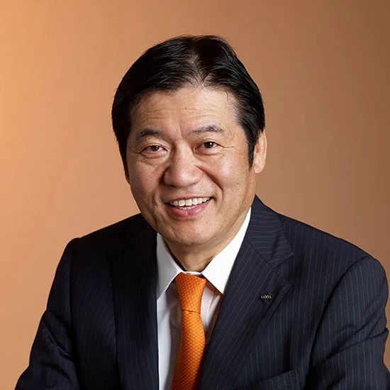 A man in a dark pinstripe suit, white shirt, and orange tie smiling in front of a beige background.