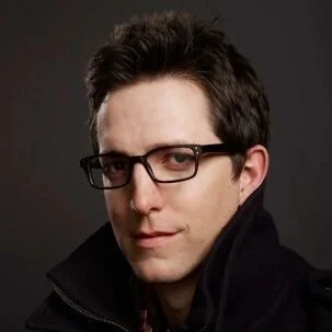 A man with dark hair wearing glasses and a black jacket, looking at the camera against a dark background.