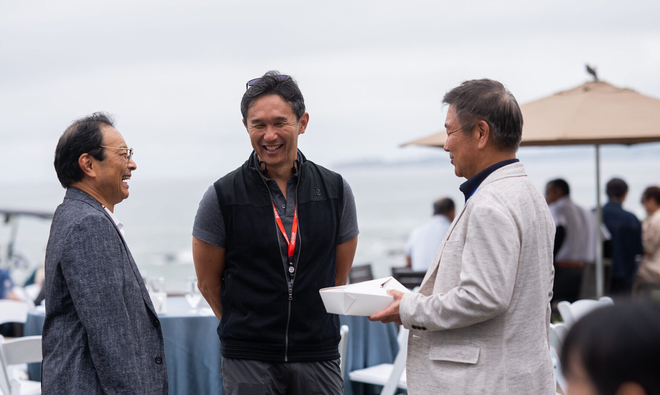 Three men are smiling and conversing outdoors, with tables and an umbrella in the background, under an overcast sky.