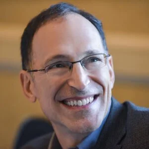 Close-up of a smiling man with glasses, short dark hair, and a suit, against a blurred warm background.