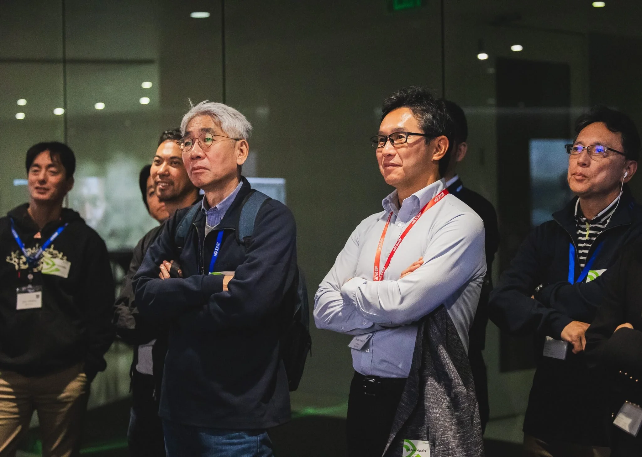 Group of five men, some wearing glasses and conference badges, standing with arms crossed and looking attentively in a dimly lit indoor setting with reflective glass walls.