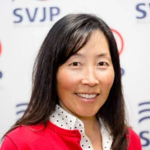 A woman with long dark hair smiling, wearing a red jacket over a white shirt with black polka dots, standing in front of a backdrop with 'SVJP' logos.