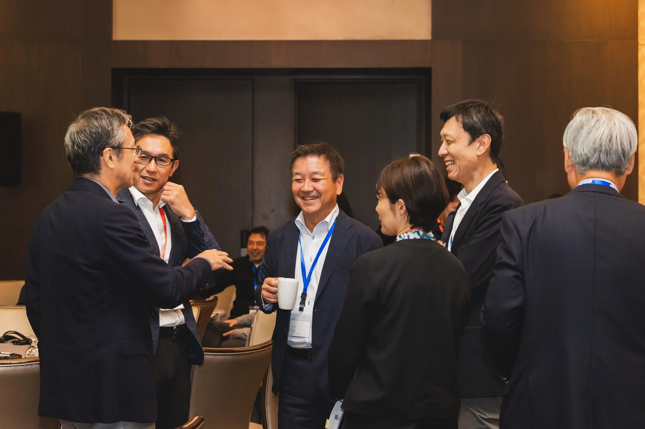 Group of six people in business attire engaging in conversation and smiling at a conference or meeting.