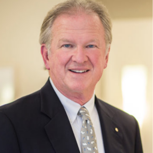 A middle-aged man with light-colored hair smiling in a professional setting, wearing a dark suit, white shirt, and patterned tie.