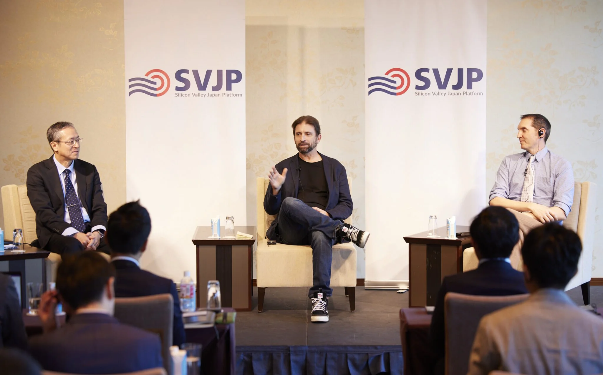 Panel discussion at an event with three men seated on stage, one speaking, two listening, with an audience in front, and banners behind with 'SVJP Silicon Valley Japan Platform' logo.