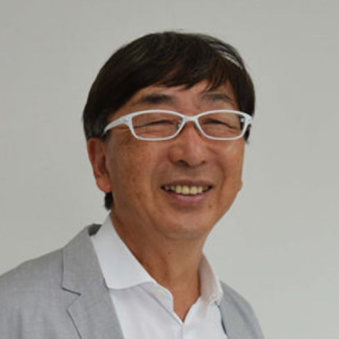 A middle-aged man with dark hair, wearing white-framed glasses, a white shirt, and a light gray blazer, smiling against a plain gray background.