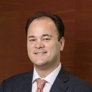 Professional portrait of a man with dark hair, wearing a dark suit and pink tie, smiling against a wooden background.