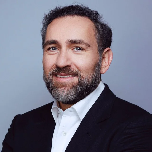 A professional headshot of a middle-aged man with a beard, wearing a black suit and white shirt, smiling against a plain gray background.