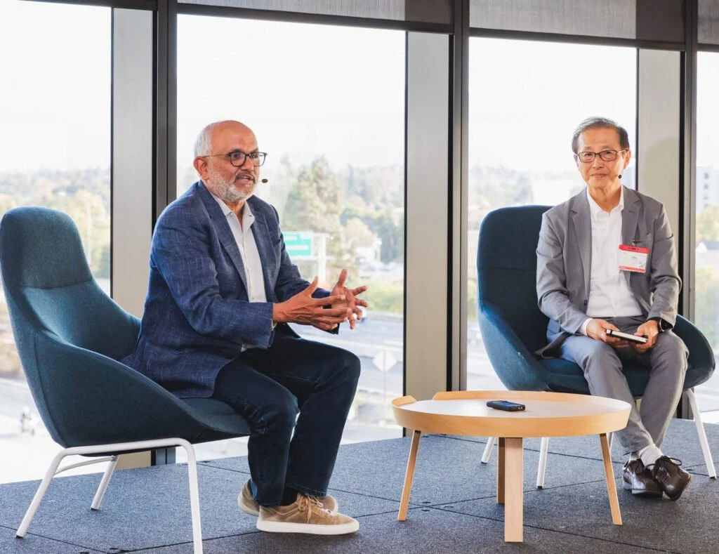 Two men sitting in modern chairs having a discussion in a bright office with large windows and a city view.
