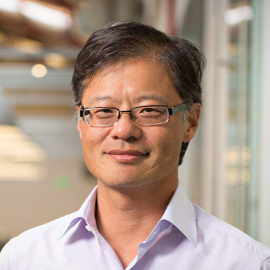 A middle-aged man wearing glasses and a white collared shirt, standing indoors near a window with blurred background.