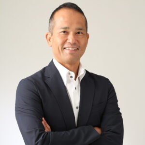 Professional man with short hair and a friendly smile, wearing a dark blazer over a white shirt, standing against a neutral background.