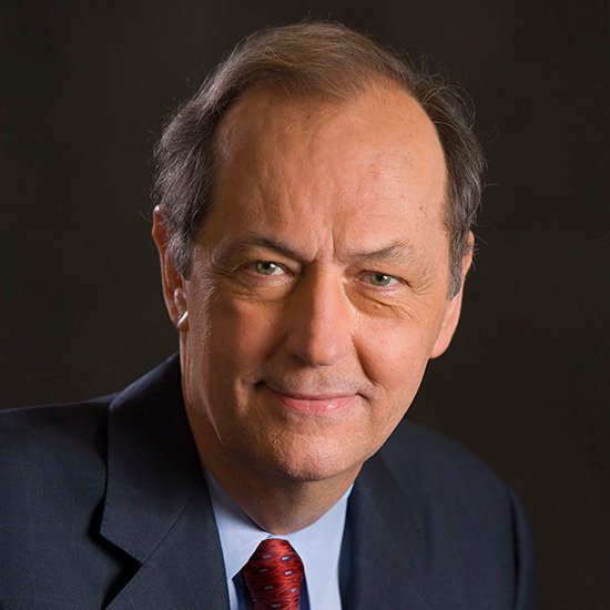 Portrait of a middle-aged man with brown hair wearing a dark suit, light blue shirt, and red tie, smiling slightly against a dark background.