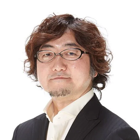 A man with wavy brown hair, wearing glasses and a black blazer over a white shirt, posing against a plain white background.