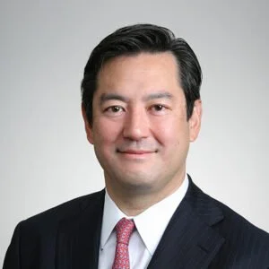 Headshot of a man with black hair, wearing a suit and tie, smiling with a neutral background.