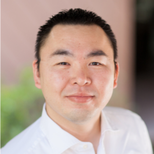 Portrait of a smiling man with short black hair, wearing a white shirt, standing outdoors with blurred greenery in the background.