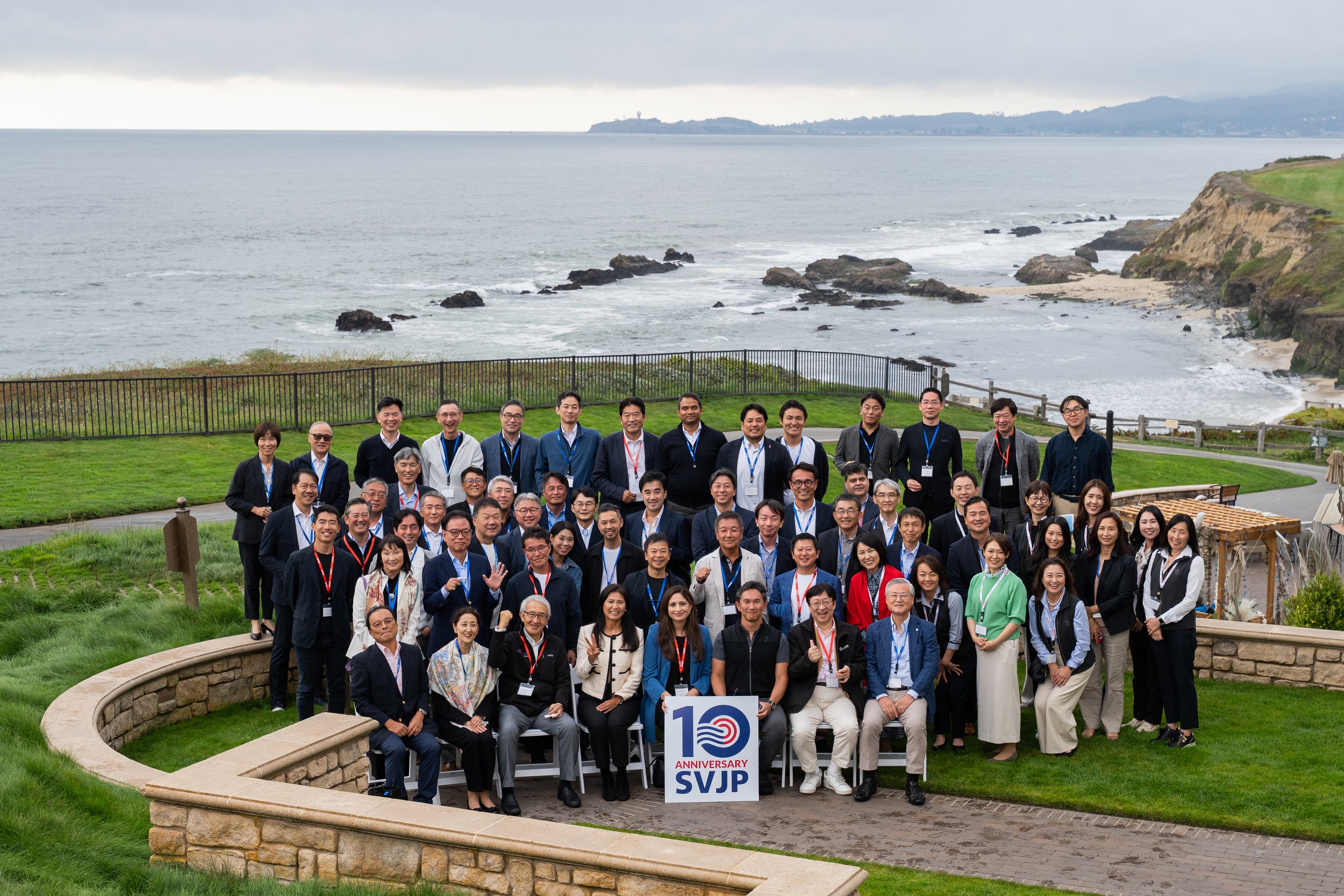 Group of diverse people gathered outdoors on a grassy area near the coast, celebrating a 10th anniversary with a sign reading '10 Anniversary SVJP'.