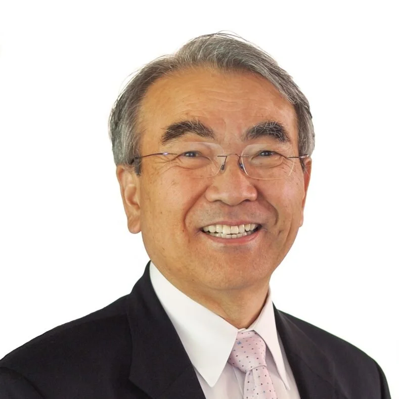 A smiling middle-aged man with glasses, wearing a dark suit, white shirt, and light pink tie, against a plain white background.
