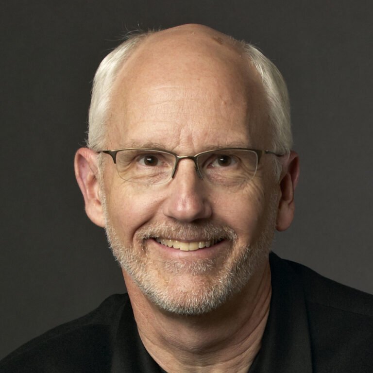 Headshot of a smiling older man with glasses and a short beard, wearing a dark shirt against a dark background.