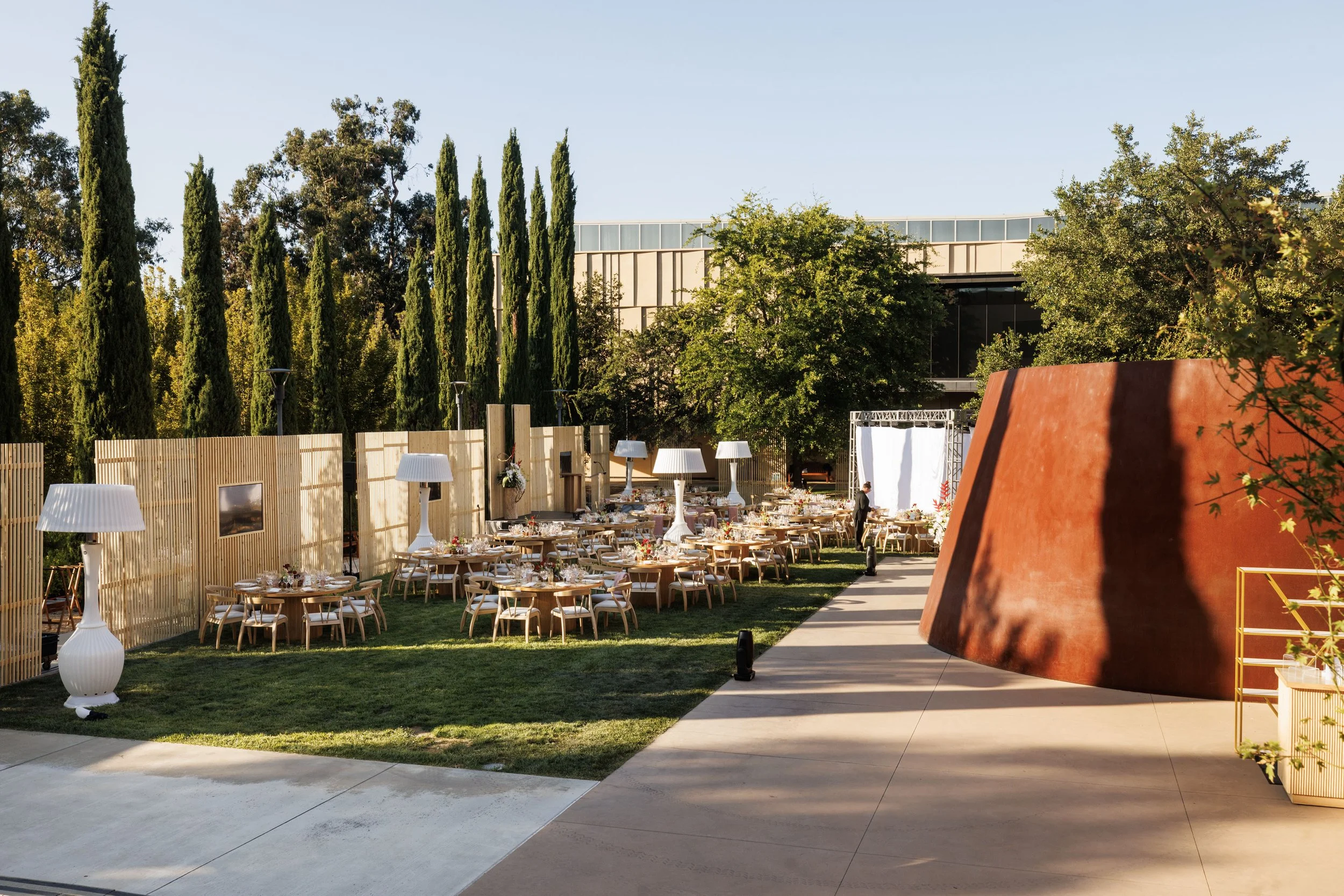 Outdoor event setup with round tables, white chairs, and white lamp fixtures, surrounded by lush green trees, a modern building in the background, and a large rust-colored sculptural piece.