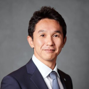 Professional man in a navy suit and light blue tie, smiling at the camera, gray background.