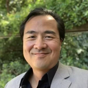 A man with dark hair and a smile, wearing a light gray blazer and black shirt, standing outdoors with green foliage in the background.